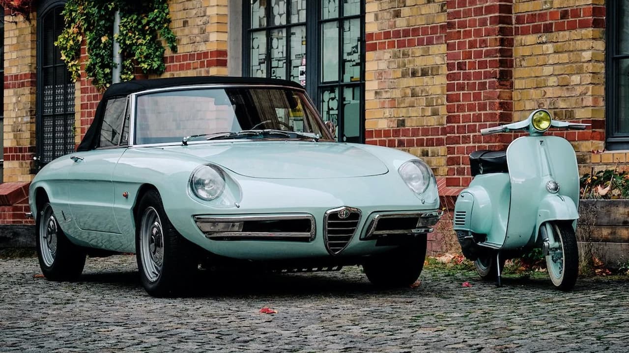 A blue Alfa Romeo Spider Duetto standing on a road with a same color scooter along